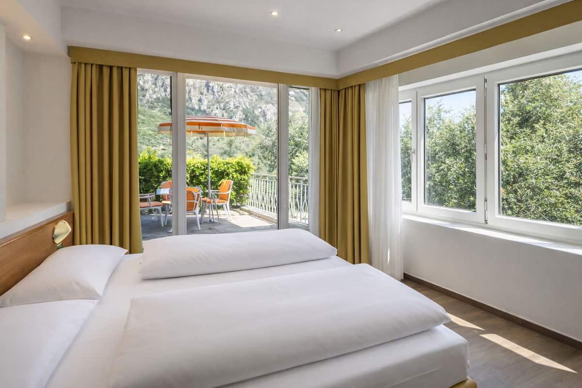Una habitación de hotel con una cama grande, dos ventanas con cortinas y un balcón con mobiliario exterior.