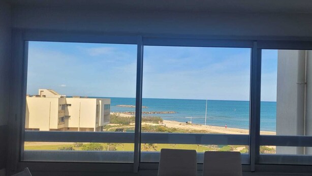 Een kamer met een groot raam met uitzicht op een strand en de oceaan, met gebouwen in de verte.