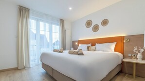 Ein großes Bett mit Holz-Kopfteil, ein Nachttisch mit Blumenvase und ein Blick ins Freie durch ein fensterbelaubtes Fenster.