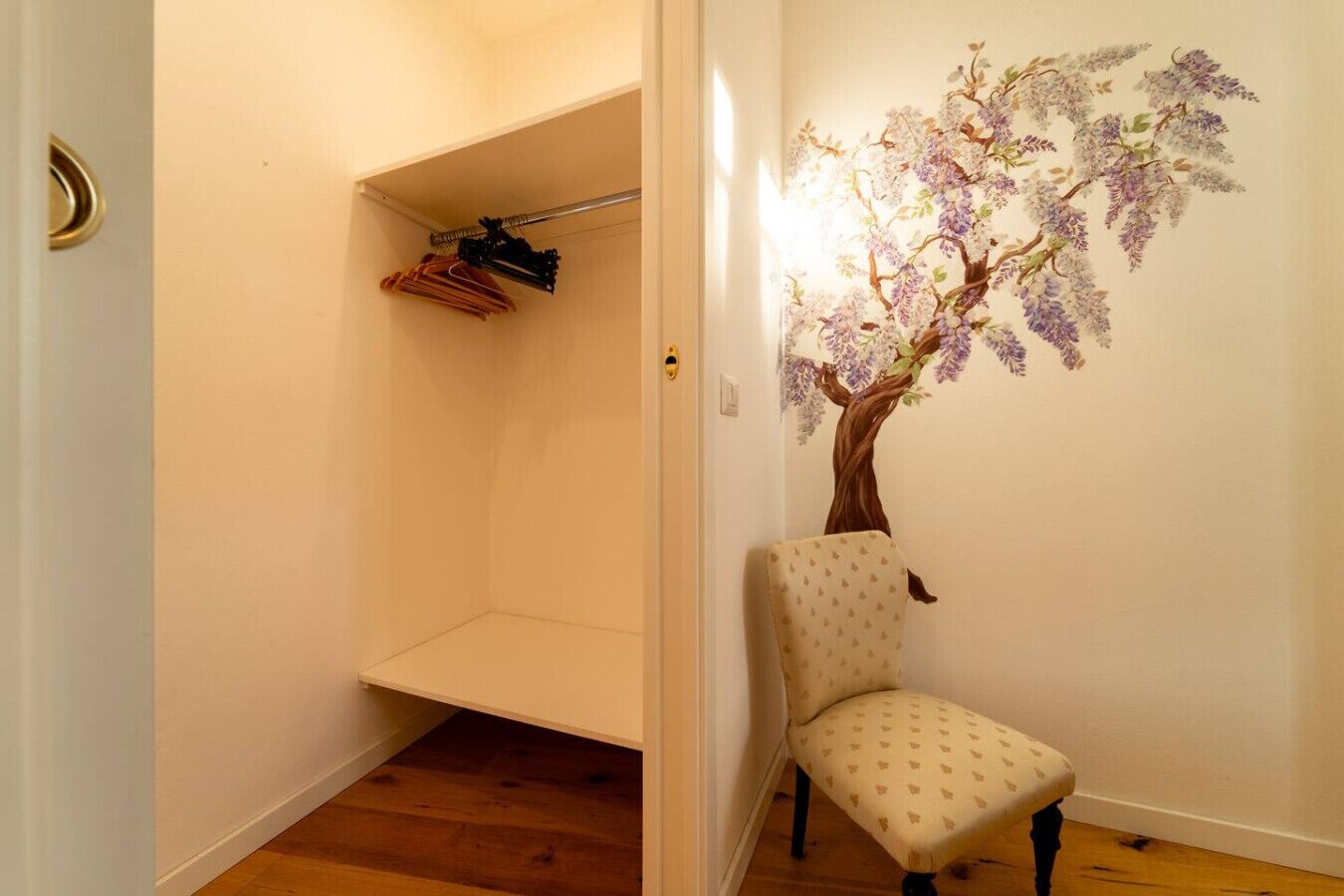 Un couloir doté d’un plancher en bois, d’une chaise, d’un porte-manteau avec des vêtements et d’une fresque murale représentant un arbre aux fleurs violettes.