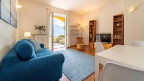 Ein modernes Wohnzimmer mit einem blauen Sofa, einem Esstisch und einem Fernseher. In der Nähe des Fensters steht ein Bücherregal und ein Sessel.