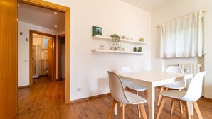 Ein moderner Essbereich mit Holzboden, Tisch und weißen Stühlen. Regale mit Dekorationselementen und ein Fenster mit Vorhängen.