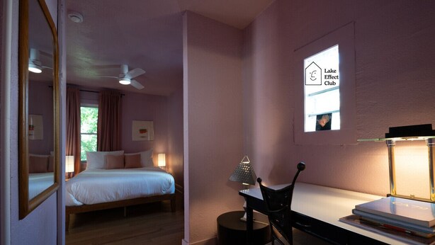 Un dormitorio con cama, escritorio, silla, lámpara, ventilador de techo y ventana con un letrero.