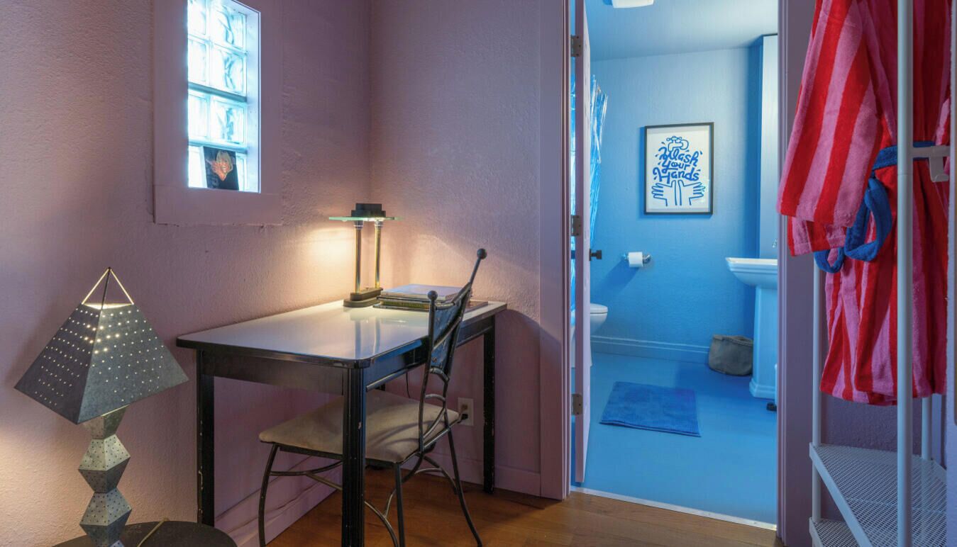 Une pièce comprenant une table, une chaise, une lampe et donnant sur une salle de bain avec un mur bleu et un panneau décoratif encadré.