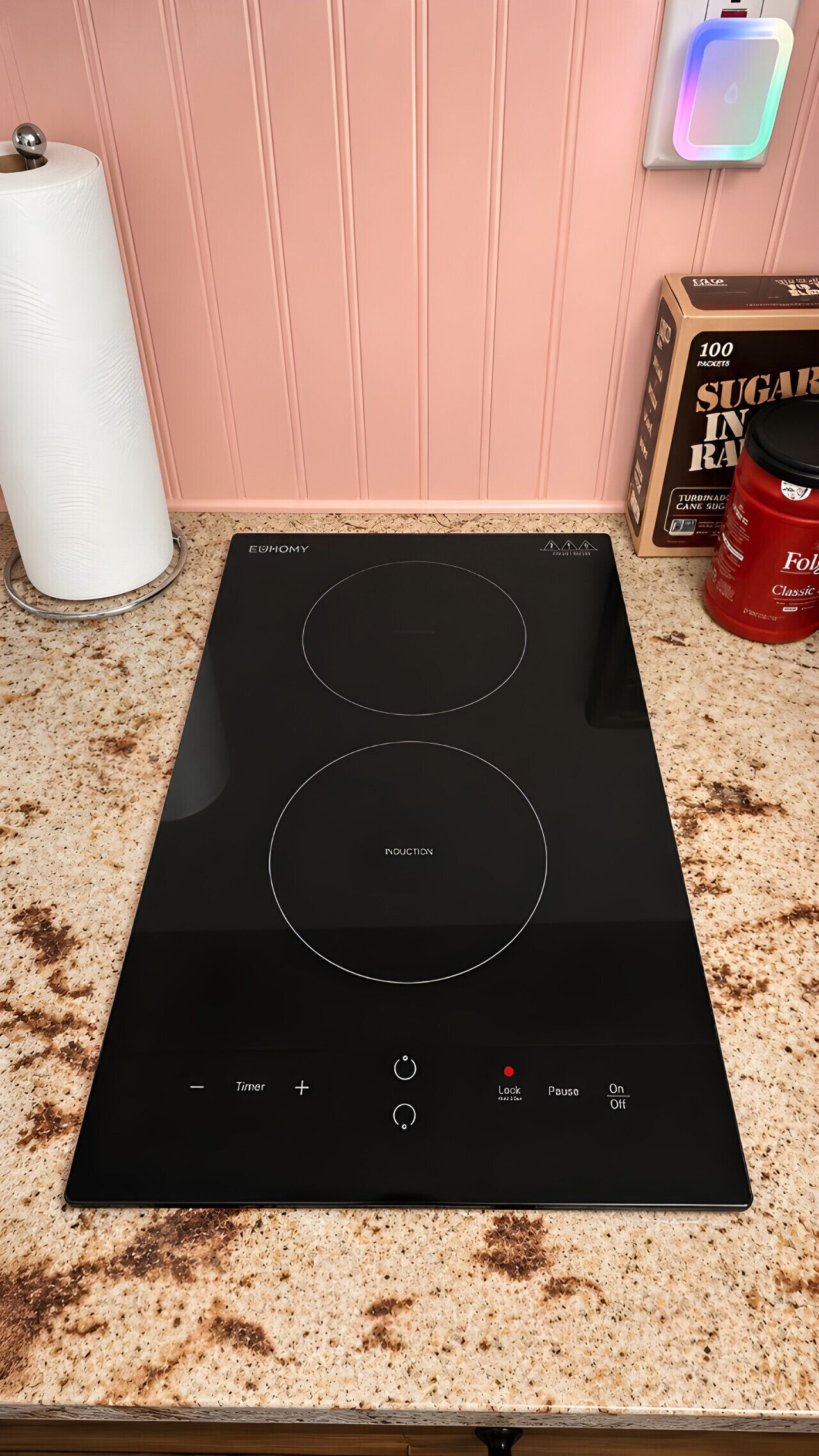 Une cuisine avec une plaque de cuisson à induction noire, un porte-essuie-tout, un paquet de sucre et un récipient rouge.