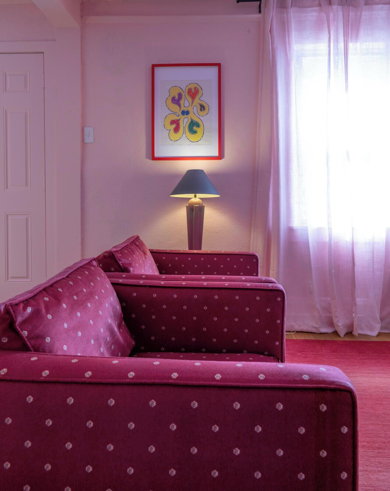 Una habitación con pared rosa, un cuadro enmarcado, una lámpara y un sofá rojo.