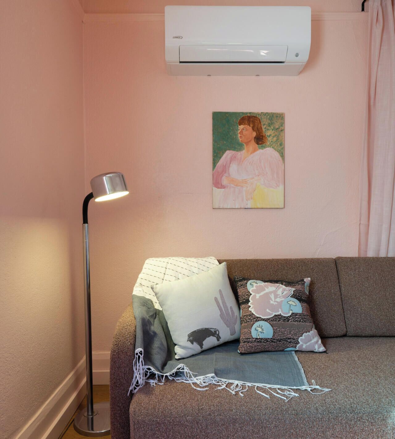 A room with a gray sofa, a white air conditioner, a floor lamp, and a painting on the wall.