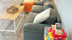 Een woonkamer met een grijze bank, een houten salontafel, een lamp en een Monopoly speldoos.