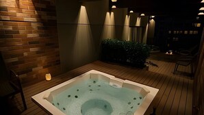 Un bain à remous avec deux verres de vin rouge, une bouteille de vin et une bougie, le tout posé sur une terrasse en bois.