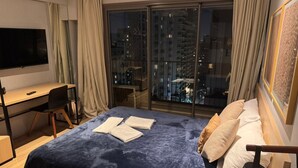 Une chambre d’hôtel équipée d’un lit, d’un bureau, d’une chaise, d’une télévision et d’une grande fenêtre donnant sur la ville.