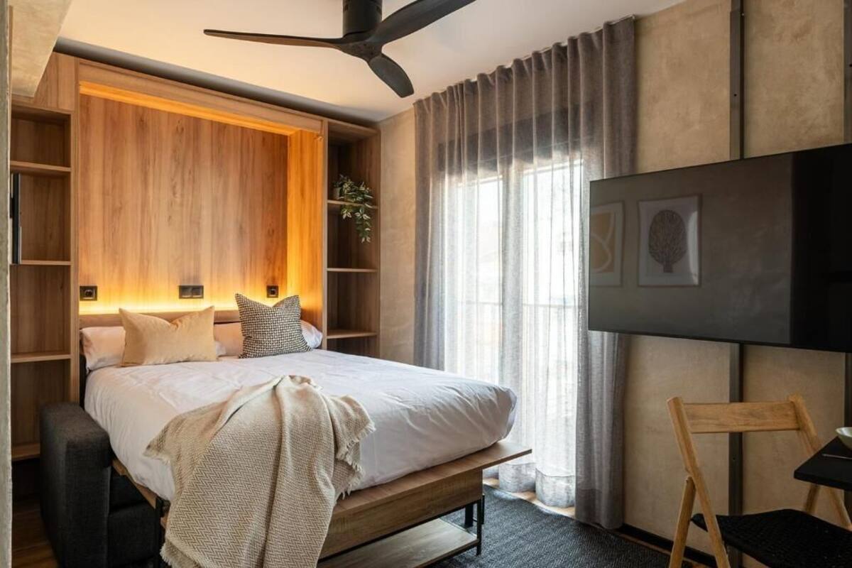 A modern hotel room with a bed, wooden headboard, built-in shelves, a TV, and a large window with curtains.