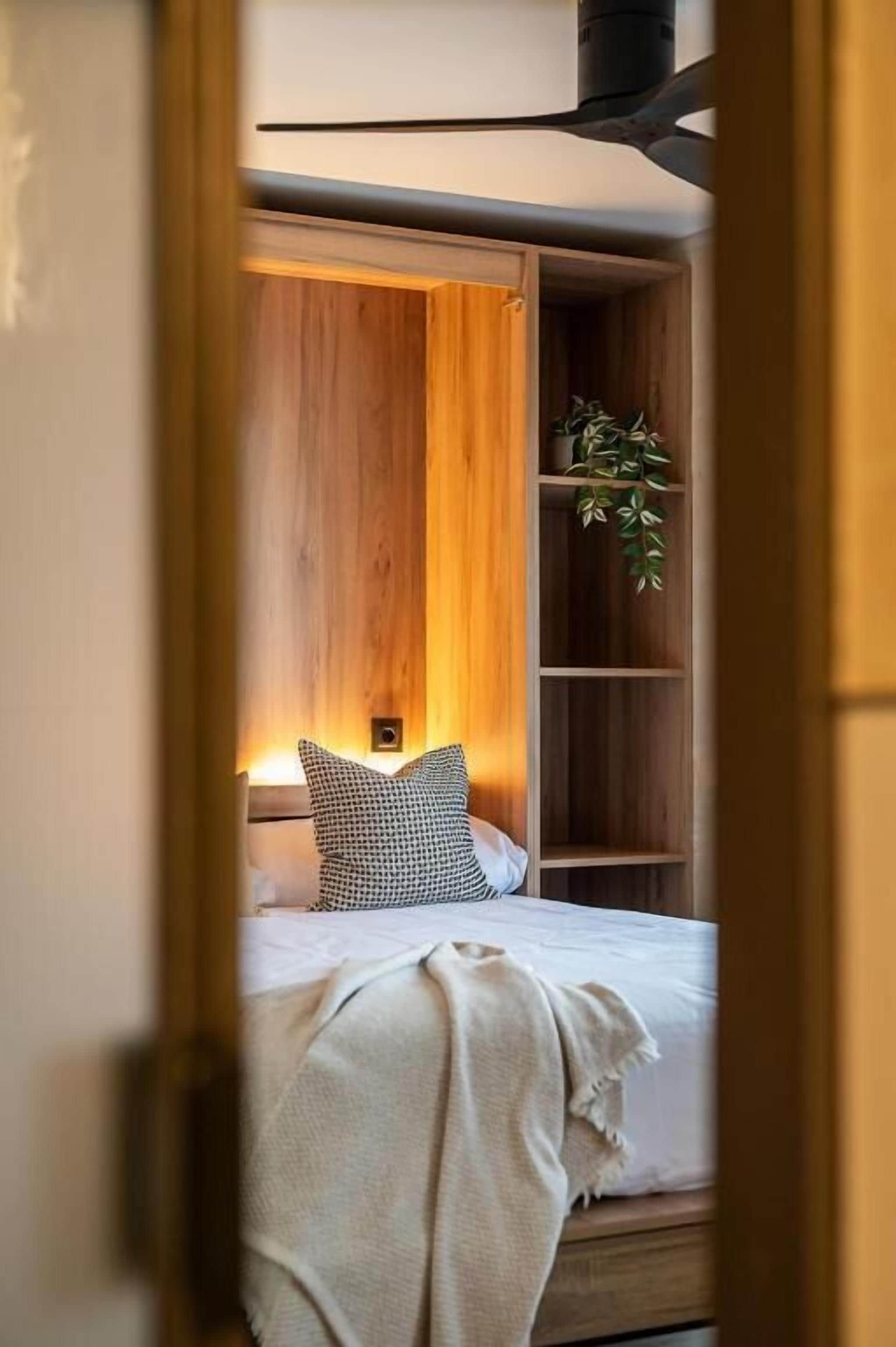 Un dormitorio con cabecera de madera, una planta en la mesita de noche, un cojín estampado y una lámpara de noche.