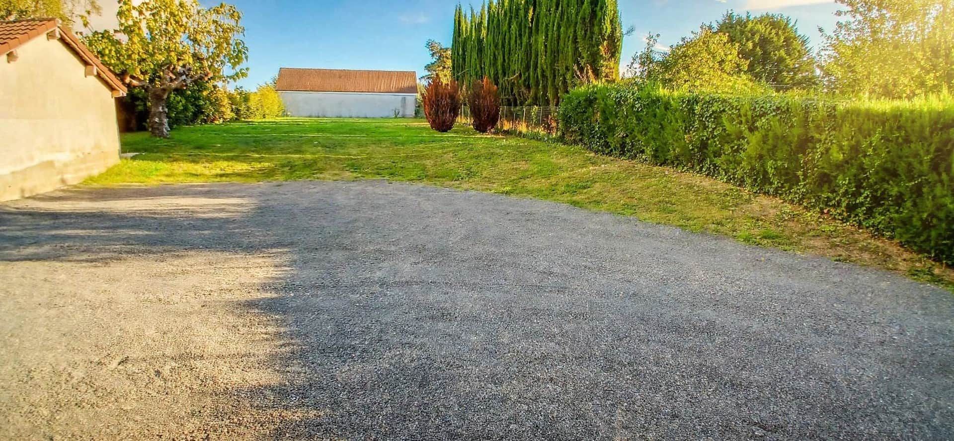 Une allée de gravier mène à un bâtiment doté d’un toit en pente, qui est entouré de verdure et d’arbres.