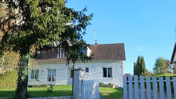 Une maison à deux étages, avec un toit recouvert de tuiles, une clôture blanche et un ciel d’un bleu limpide.