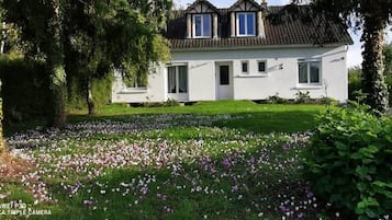 Une maison blanche avec un toit en pente, entourée d’un jardin où les fleurs sont en pleine floraison.