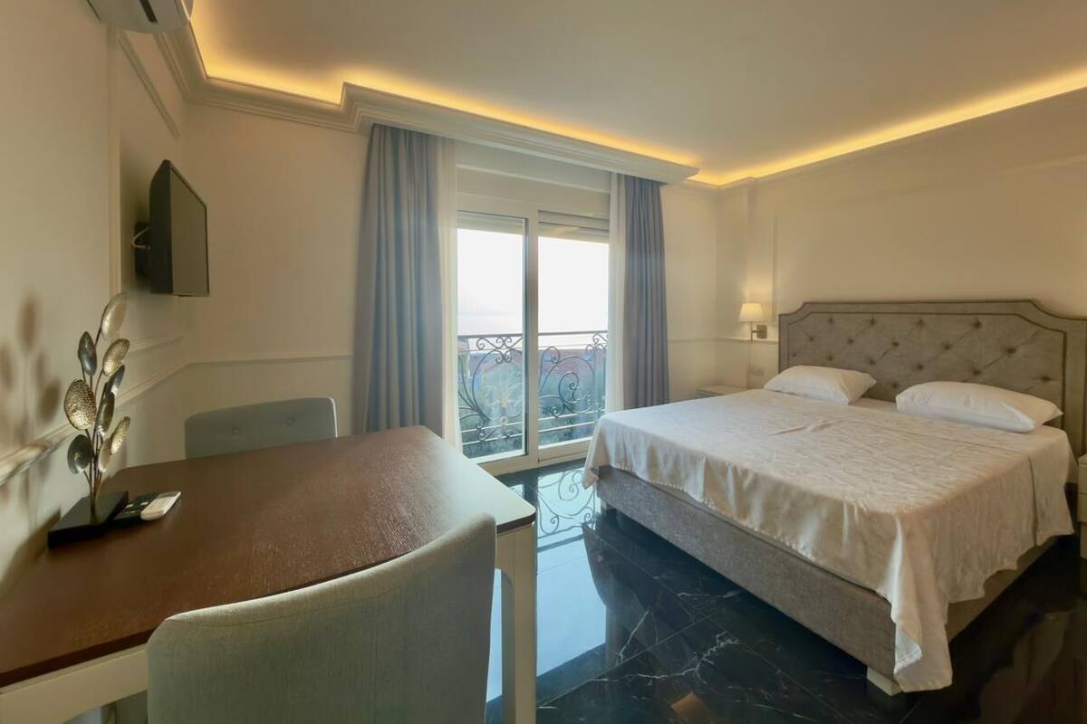 A hotel room with a bed, a desk, and a chair. There is a window with curtains and a balcony.