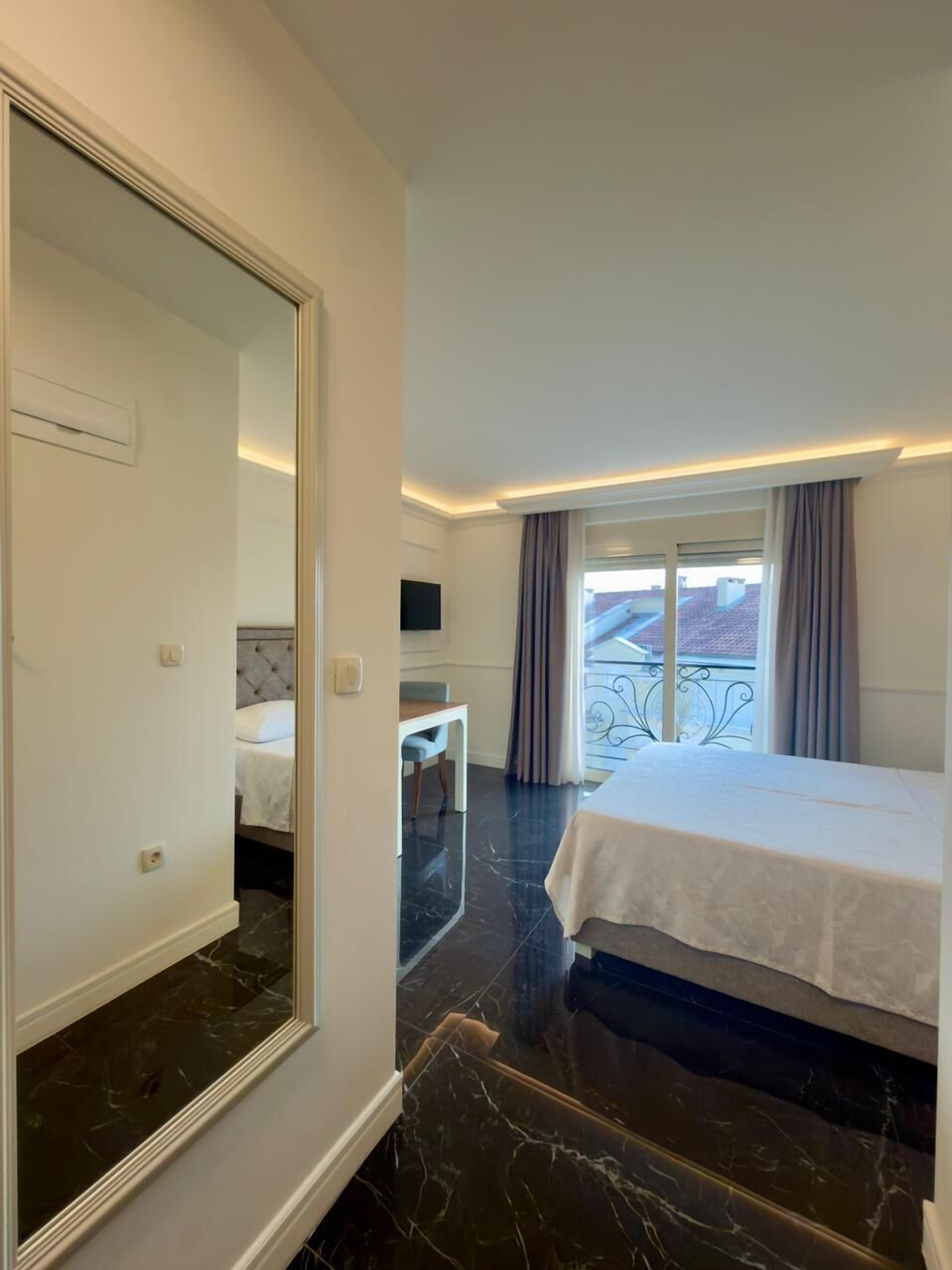A hotel room with a large mirror, a bed, a small desk, and a window with curtains.