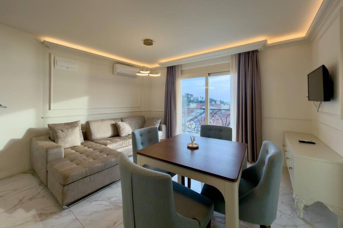 A modern living room with a sofa, dining table, and chairs. There is a TV on the wall and a window with curtains.