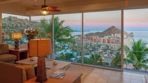 A spacious living room with a large window offering a view of a coastal cityscape, a ceiling fan, and a table with a lamp and decorative items. - Pedregal Penthouse with Breathtaking Views (Cabo San Lucas)