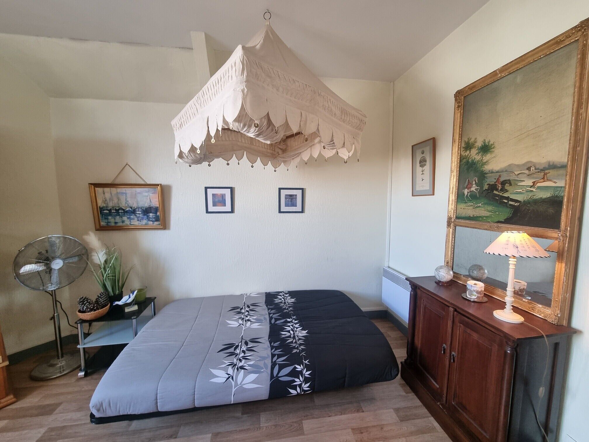 A bedroom with a bed, a fan, a nightstand, a lamp, and a painting on the wall.