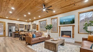 A spacious living area with a fireplace, wooden ceiling, and large windows. - New! 6BR Luxe Pool Cabin*Hot Tub*Games*Fireplace (Sevierville)