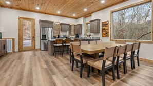 A modern kitchen with wooden cabinets, a stainless steel refrigerator, and a dining area with a wooden table and chairs. - New! 6BR Luxe Pool Cabin*Hot Tub*Games*Fireplace (Sevierville)