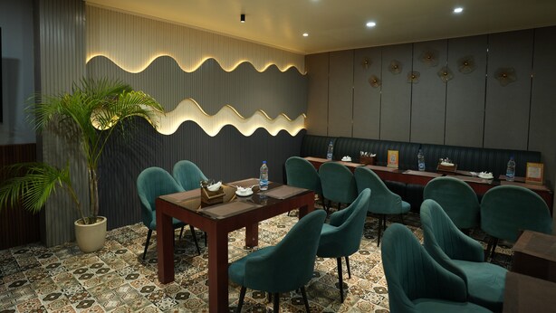 Quarto de hotel com área de refeições, destacando cadeiras em tons de verde-água, uma mesa de madeira e uma parede decorativa com padrão ondulado.