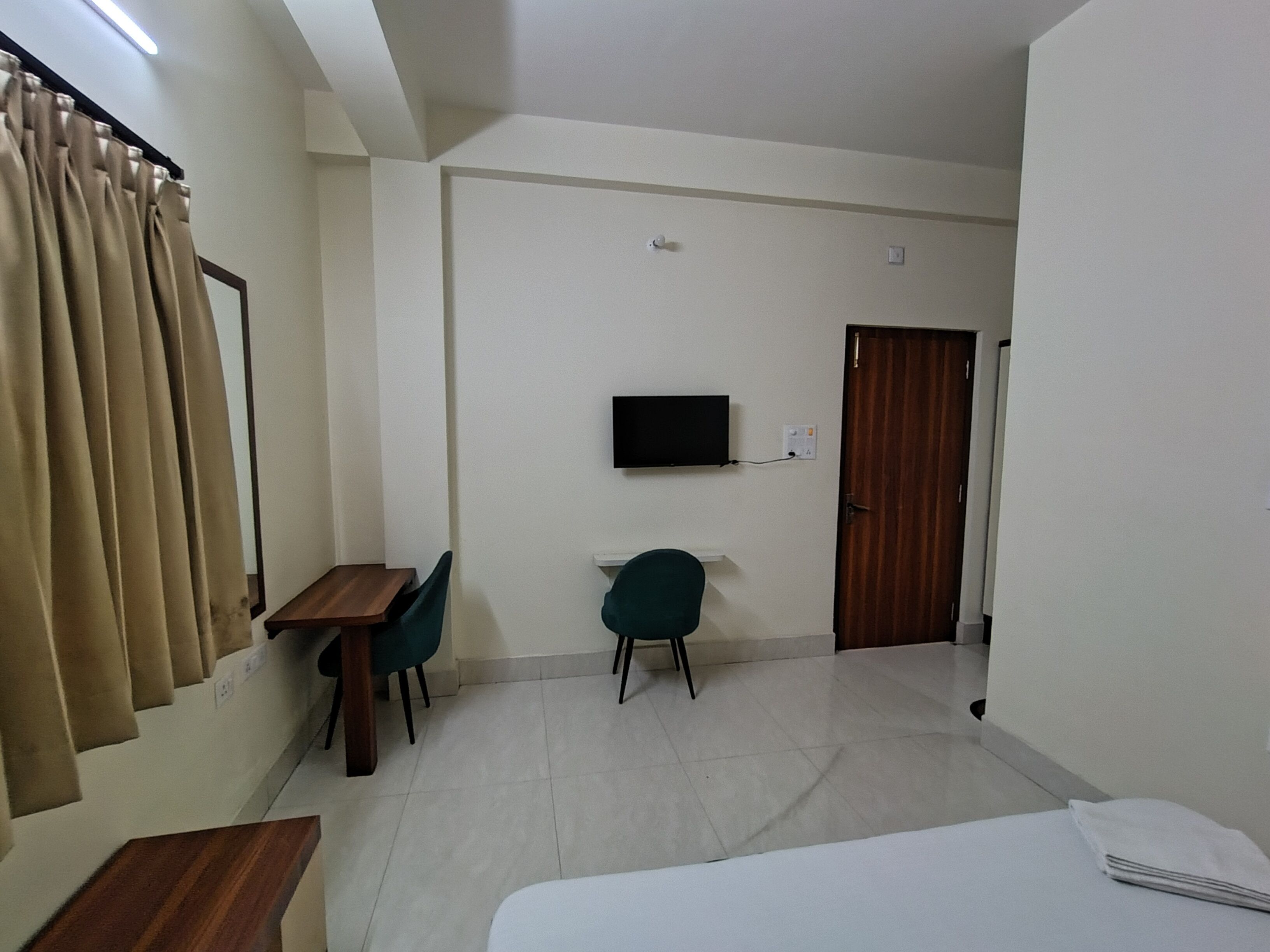 A hotel room with a bed, a desk, two chairs, a television mounted on the wall, and a door.