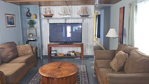 Uma sala de estar com dois sofás, uma mesa de centro, uma TV e objetos decorativos como réplicas de navios e um quadro emoldurado.