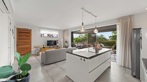 A modern kitchen with a central island, a dining area with a table and chairs, a sofa, and a large window with a view of greenery. - Apartment Casa Sweet, Jan Sofat, Pool & Tropical Garden (Willemstad)