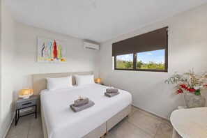A bedroom with a bed, bedside lamps, a window with a view, an air conditioner, and a painting on the wall. - Apartment Casa Sweet, Jan Sofat, Pool & Tropical Garden (Willemstad)