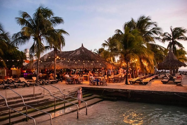 A beachside restaurant with outdoor seating, palm trees, and a thatched-roof structure.
