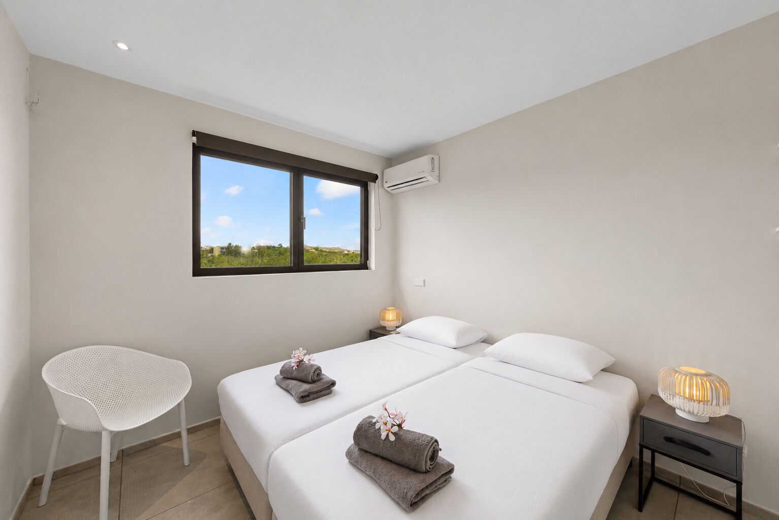 A bedroom with two beds, a chair, a bedside table with a lamp, and a window with a view.