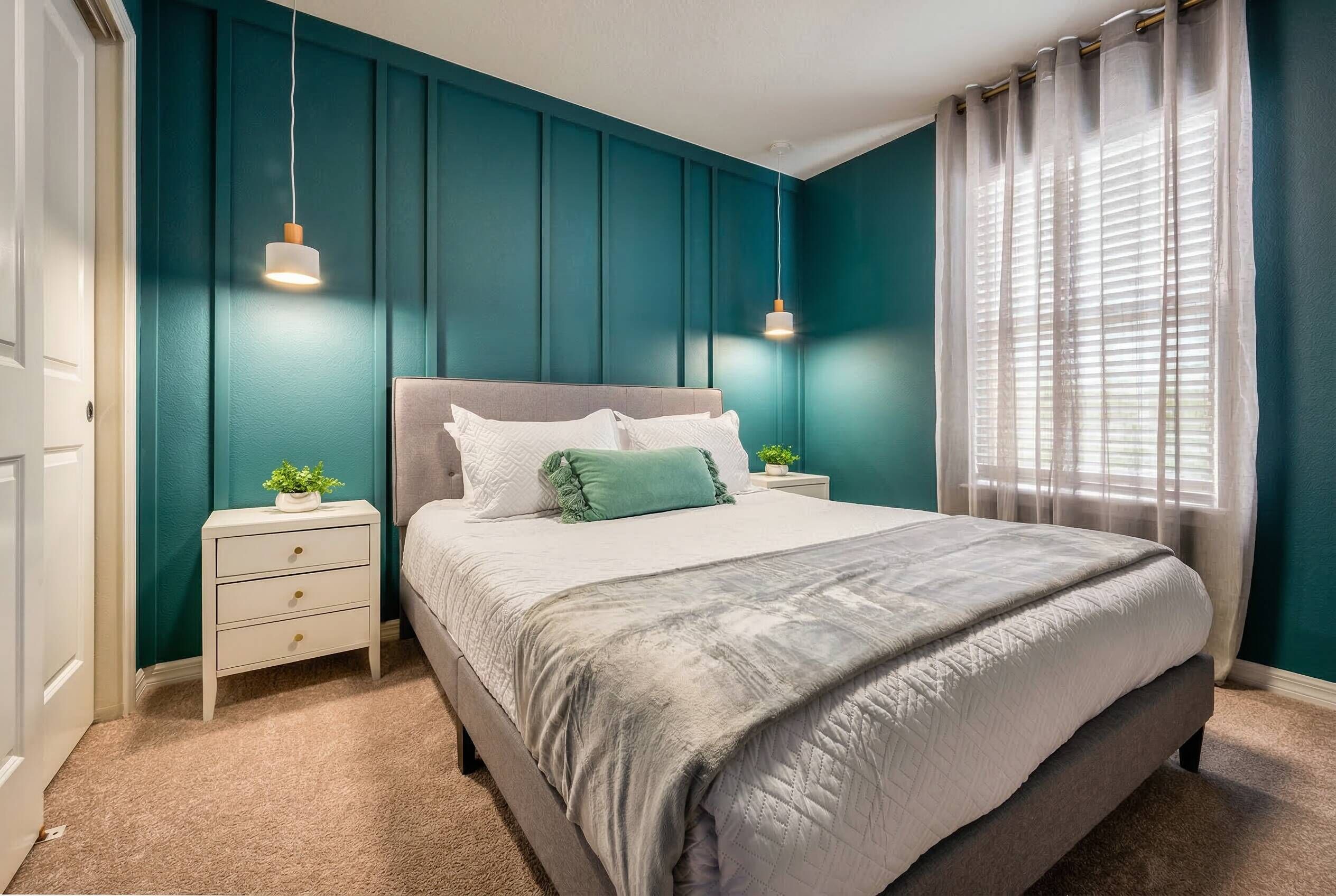 A bedroom with teal walls, a bed with a grey headboard, two bedside tables, and a window with sheer curtains.