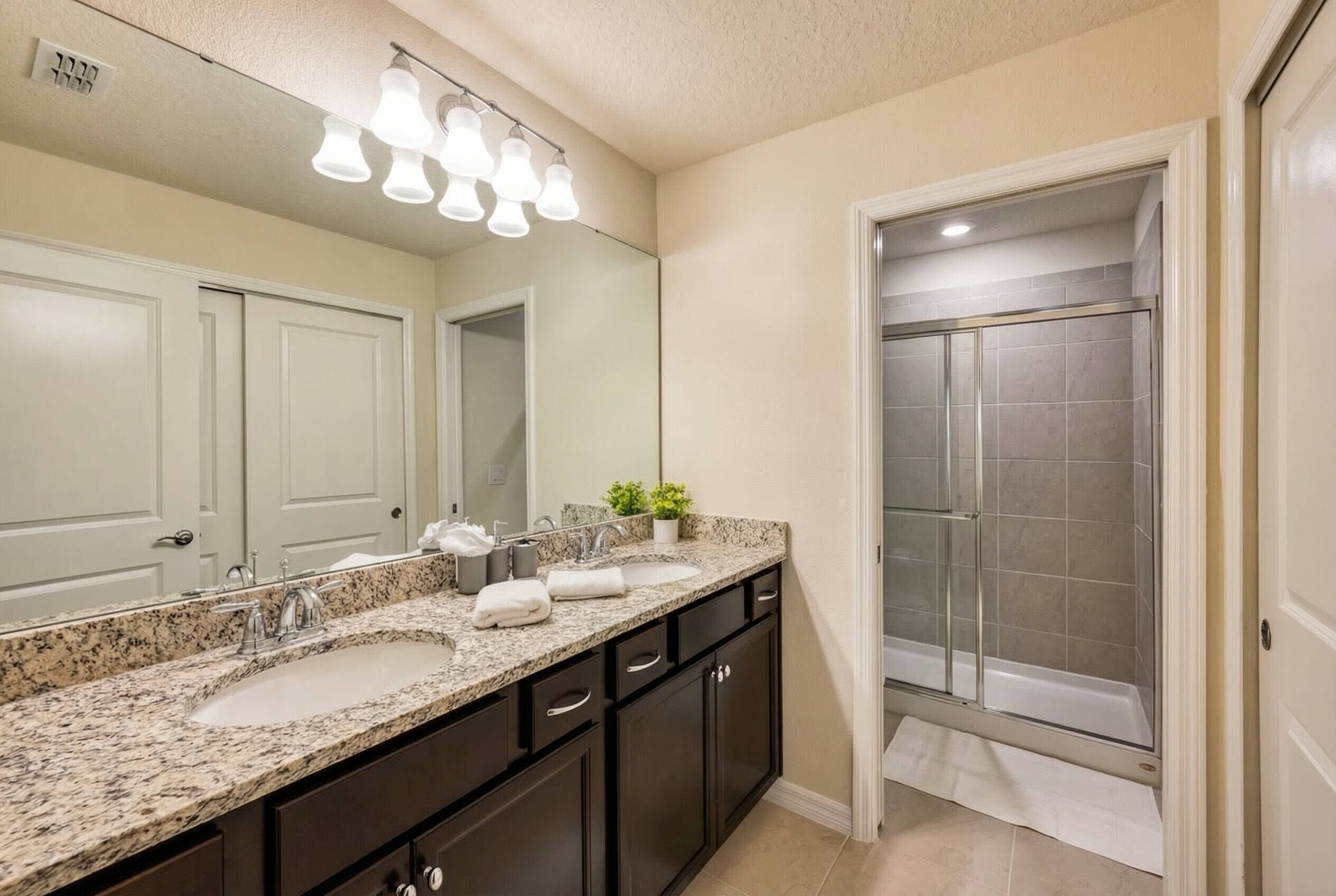 A bathroom with a granite countertop sink, dark cabinets, a large mirror, and a glass shower enclosure.