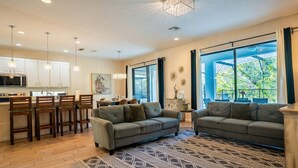 A modern living room with a sofa, chairs, a patterned rug, and a kitchen area with white cabinets and pendant lights. - 6BR Family Home w/ Pool Near Disney (Kissimmee)