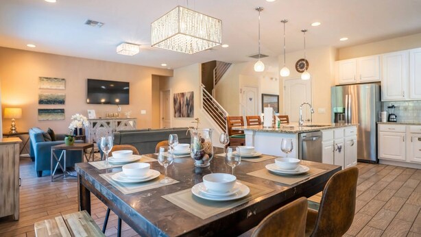 A modern kitchen with white cabinets, a dining area with a wooden table set for four, and a staircase leading to another level. - 6BR Family Home w/ Pool Near Disney (Kissimmee)
