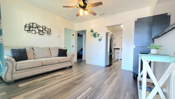 A spacious living room with a beige sofa, wooden flooring, a ceiling fan, and a kitchen area visible through an open doorway.