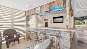 Un área de bar al aire libre con mobiliario de mimbre, una barra de piedra y un televisor montado en la pared.