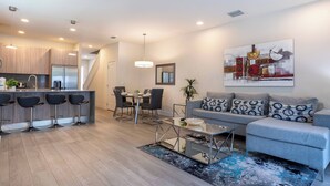 Un appartement moderne comprenant une cuisine, un coin repas et un salon. Il est doté d’un bar avec des tabourets, d’un canapé agrémenté de coussins à motifs et d’un grand tableau abstrait accroché au mur.