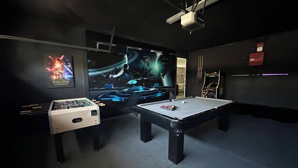 A room with a pool table, a foosball table, a large screen displaying a space-themed movie, and a projector mounted on the ceiling. - 9BR Pool & Spa Home Near Disney | Encore (Kissimmee)
