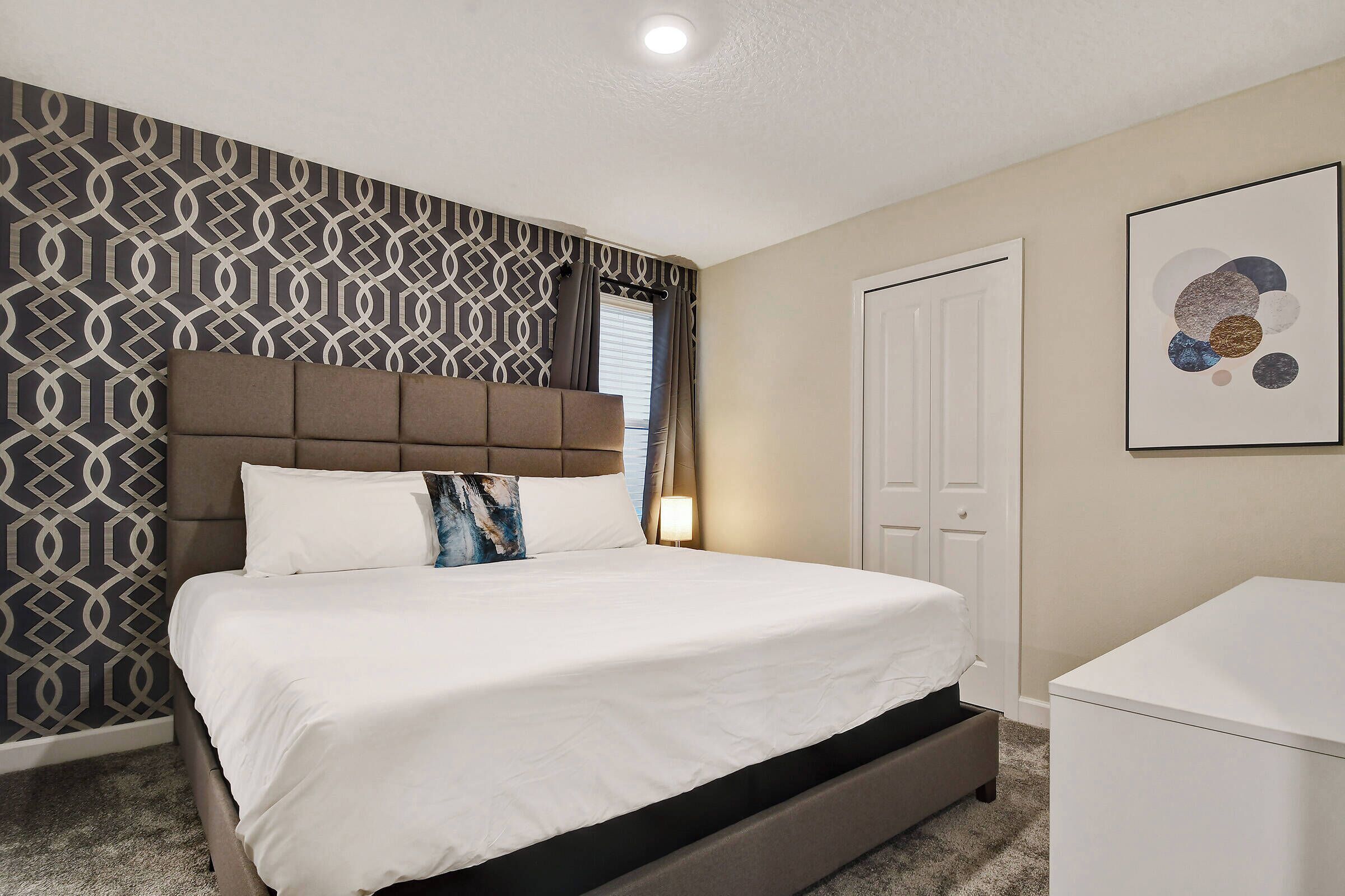 A bedroom with a bed, a headboard, a bedside table, a wardrobe, and a wall with a geometric pattern.