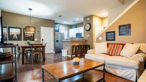 A living room with a sofa, coffee table, dining table, and chairs. There is a kitchen in the background with a refrigerator, microwave, and cabinets. - Home in West Lucaya | 4BR Near Disney (Kissimmee)