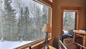 A cozy room with a large window offering a snowy forest view, a wooden table with a lamp, and a cushioned chair. - Large 5BR Ski-In/Ski-Out | Sauna | Sleeps 12 (Lutsen)