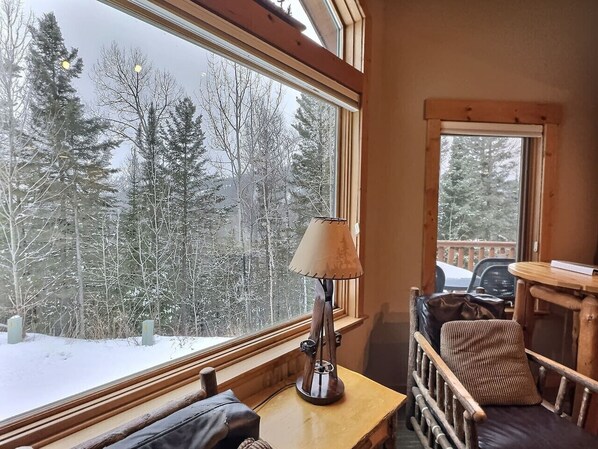 Una habitación acogedora con un ventanal grande que ofrece vistas a un bosque nevado, una mesa de madera con una lámpara y un sillón con cojines.