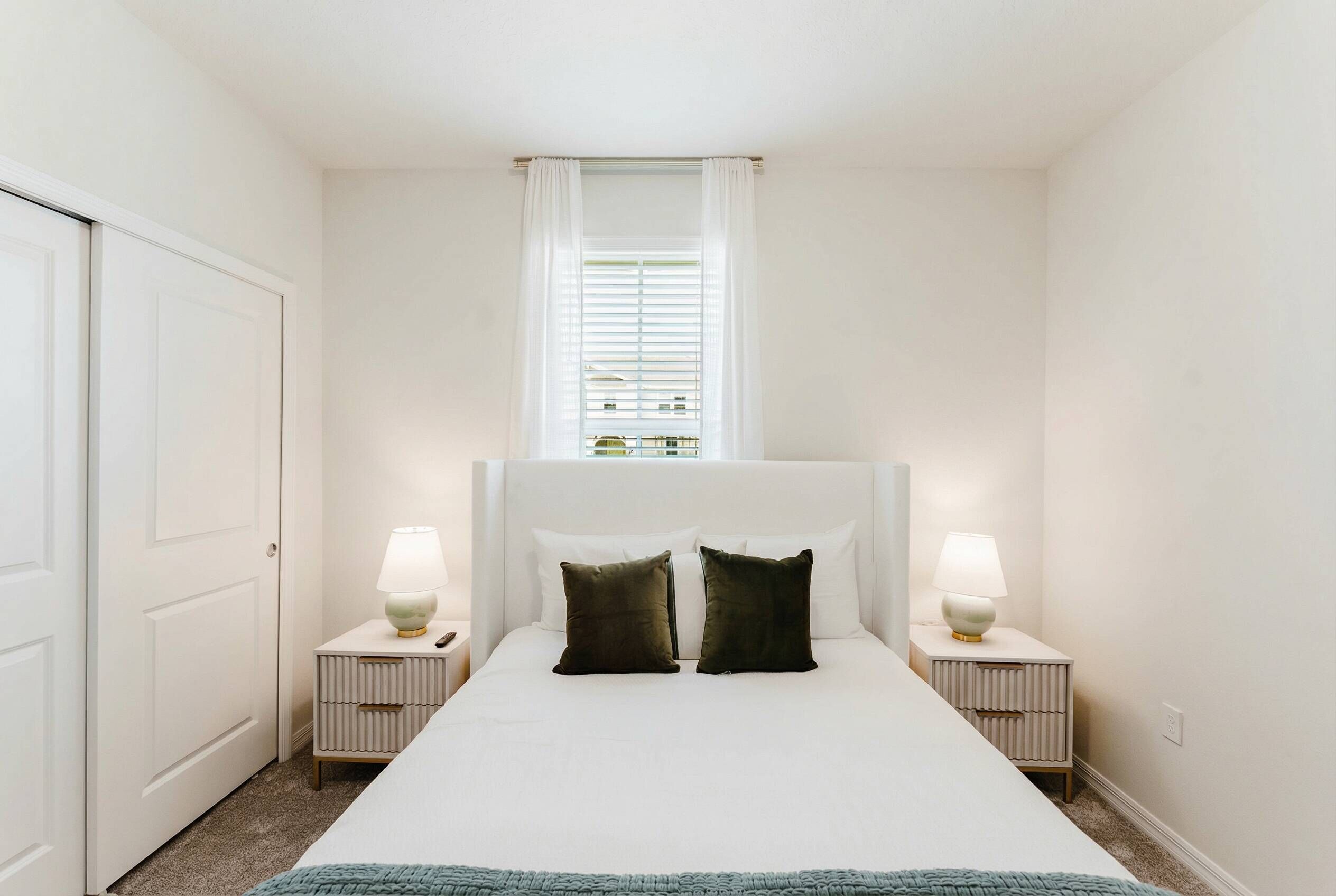Une chambre à coucher avec un lit, deux tables de chevet avec des lampes, une fenêtre avec des stores et une porte.