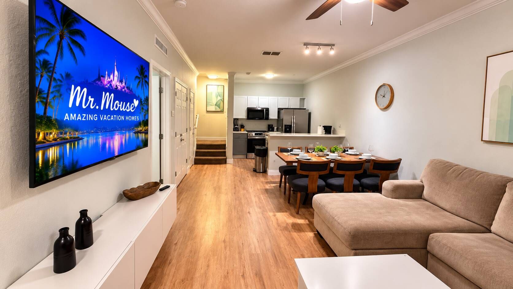 Ein modernes Wohnzimmer mit einem Flachbildfernseher, einem beigefarbenen Sofa, einem Essbereich mit einem Holztisch und Stühlen sowie einer Küche im Hintergrund.