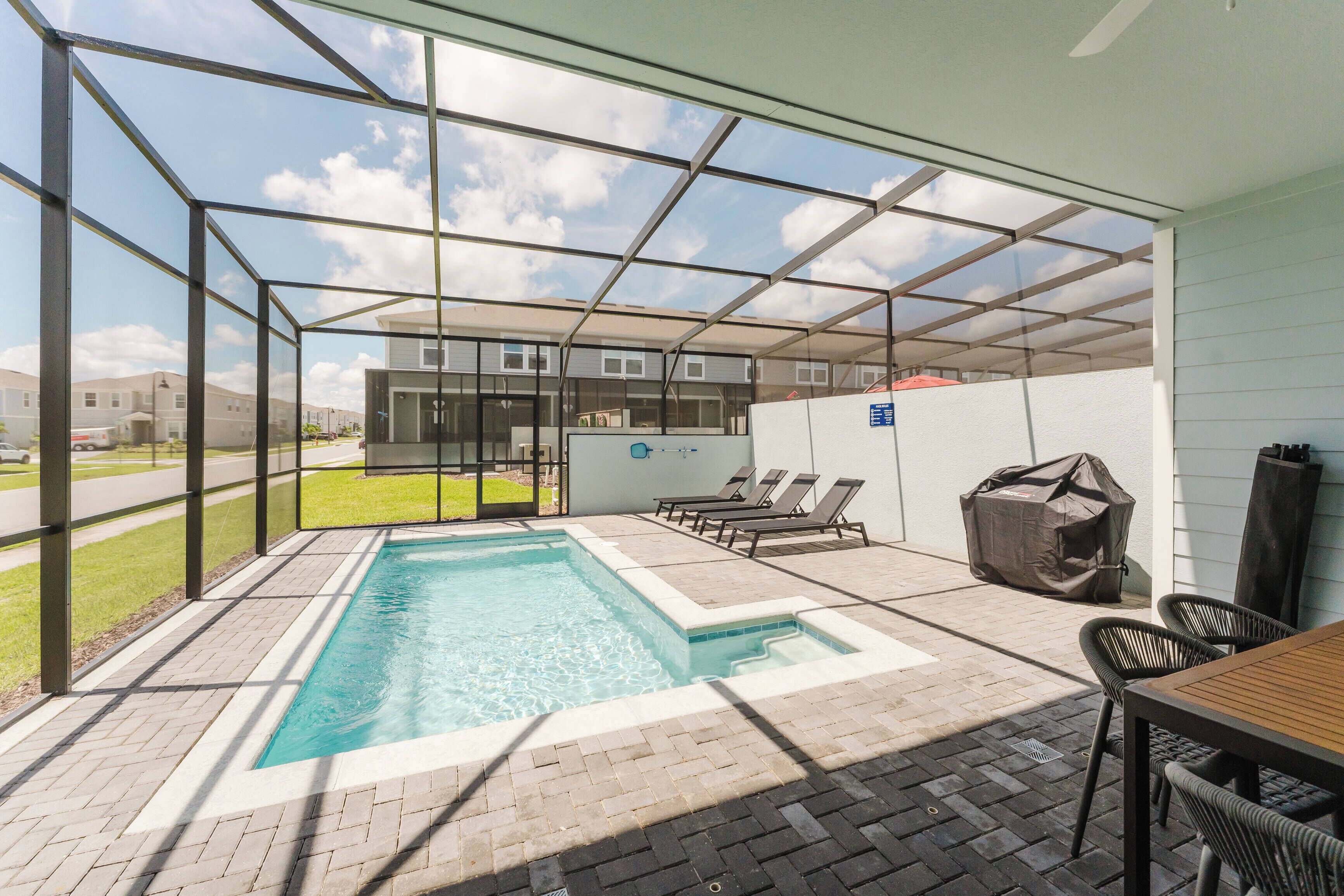 A covered outdoor swimming pool with lounge chairs and a table, surrounded by a brick patio.
