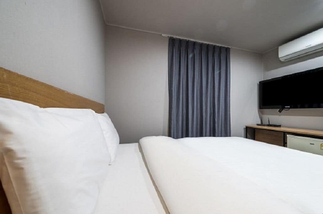 A hotel room with a bed, a desk, a flat-screen TV, and an air conditioning unit.