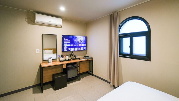 A hotel room with a bed, a desk with a computer monitor, a small fridge, a mirror, and a window with curtains. - Hotel remi (Seoul)